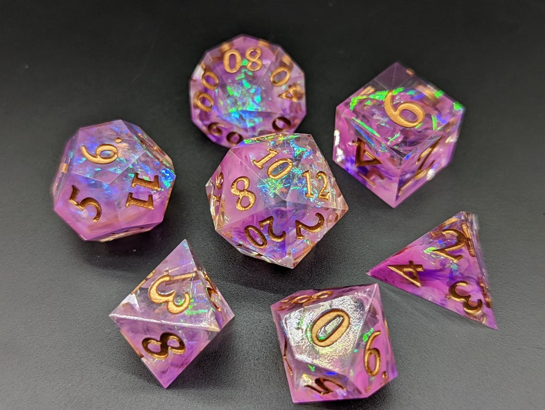May include: Set of seven iridescent purple dice with gold numbers. The dice are made of clear resin with a purple tint and have a glittery finish. The numbers are printed in gold.