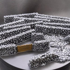 Puede incluir: Un conjunto de muros de piedra en miniatura y una puerta, ideal para juegos de mesa o modelismo. Los muros son grises con detalles de piedras individuales, y la puerta es de color dorado contrastante. Las piezas est&aacute;n dispuestas sobre una superficie gris.
