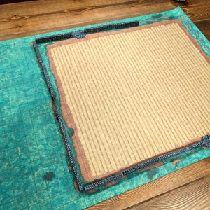 Puede incluir: Una alfombrilla de juego de mesa con un dise&ntilde;o de mapa detallado. La alfombrilla tiene un &aacute;rea central beige texturizada, una secci&oacute;n con patr&oacute;n de cuadr&iacute;cula azul-verdosa y un borde con un dise&ntilde;o de muro de piedra. La est&eacute;tica general recuerda un entorno de fantas&iacute;a.