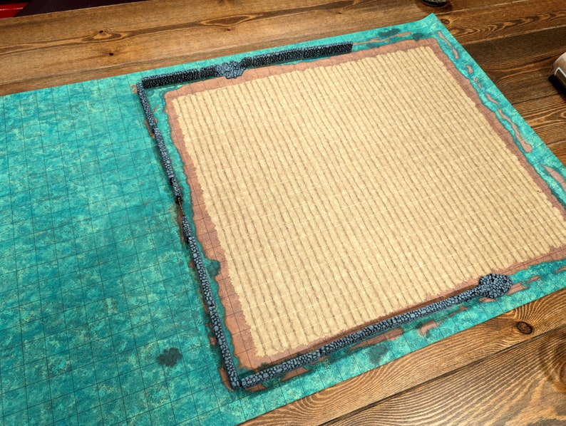 Puede incluir: Una alfombrilla de juego de mesa con un dise&ntilde;o de mapa detallado. La alfombrilla tiene un &aacute;rea central beige texturizada, una secci&oacute;n con patr&oacute;n de cuadr&iacute;cula azul-verdosa y un borde con un dise&ntilde;o de muro de piedra. La est&eacute;tica general recuerda un entorno de fantas&iacute;a.