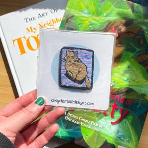 Puede incluir: Un parche de gato bordado de color marrón con un fondo a rayas azul y morado. El parche está en una tarjeta blanca con el texto "amycharlottedesigns.com" y "Una película de Studio Ghibli dirigida por Hiromasa Yonebayashi".