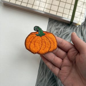 May include: Embroidered orange pumpkin patch with a green stem and brown outline. The patch is held in a hand, with a notebook and a grey, fuzzy surface in the background. The pumpkin is a seasonal item.