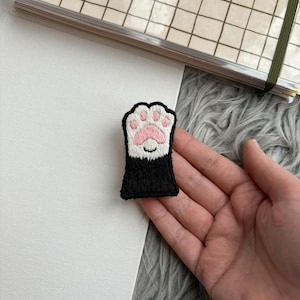 Puede incluir: Parche bordado con un diseño de pata de gato. El parche tiene una base negra con una almohadilla rosa y detalles blancos. El parche es rectangular y tiene un borde cosido. El parche se sostiene en una mano.