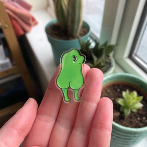 May include: A green cartoon character with a smiling face and large eyes. The character is standing with its legs spread apart and its arms raised above its head. The character is made of clear acrylic and has a glossy finish.