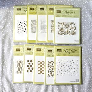 May include: A collection of Stampin' Up! Big Shot embossing folders in various designs. The folders feature patterns such as stars, wood grain, floral, and geometric shapes. The packaging is light green and white.
