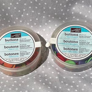 May include: Four clear plastic containers of buttons with white labels and blue text. The containers are filled with buttons in various colors, including blue, red, green, and pastel shades. The labels read "Buttons" and include the button color names.