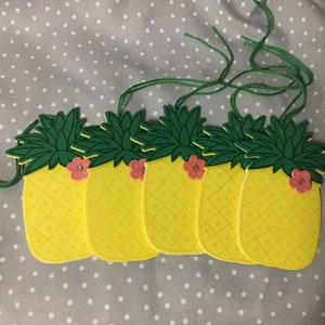 May include: A set of six yellow pineapple-shaped paper ornaments with green tops and pink flowers. Each ornament has a green string attached for hanging.