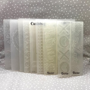 May include: A collection of clear, rectangular embossing folders with various designs, including floral patterns, hearts, and the words "Valentine's Day," "XOXO," and "To my Love." The brand name "Darice" is visible on some folders.