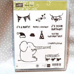 May include: Clear plastic case containing a set of rubber stamps with various celebratory designs and phrases. Includes an elephant, banners, hearts, and the text "It's a Party!" and "Merry Christmas!"