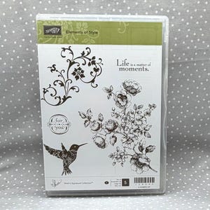 May include: A clear plastic stamp storage case containing a sheet with black ink illustrations of a hummingbird, floral branches, a flourish, and the text Life is a matter of moments and for you. The Stampin Up logo is visible at the top left.