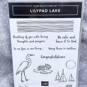 May include: Clear-mount stamp set titled "LILYPAD LAKE" with various line art designs. Includes a heron, lily pads, sailboats, and phrases like "Thinking of you with loving thoughts and prayers." The set also includes the word "Congratulations".