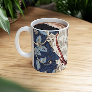 Puede incluir: Taza de café de cerámica blanca llena de café oscuro. La taza presenta un diseño floral con flores blancas, detalles rojos y hojas azules sobre un fondo azul oscuro. La taza tiene un asa blanca.