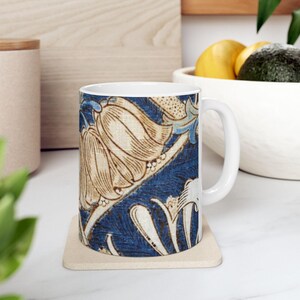 Puede incluir: Taza de cerámica blanca con un diseño floral azul y beige. El diseño incluye flores y hojas estilizadas sobre un fondo azul oscuro. La taza está sobre un posavasos beige, con un asa blanca.