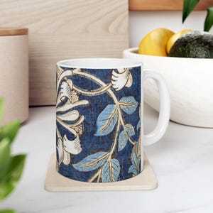Puede incluir: Taza de cerámica blanca con un estampado floral azul con hojas azul claro y detalles crema. La taza está sobre un posavasos beige. El diseño recuerda a las ilustraciones botánicas vintage.