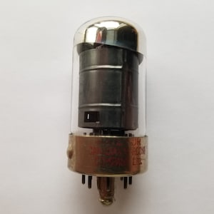 Peut inclure: Un tube à vide vintage avec une enveloppe en verre transparent et une grille en métal noir. Le tube a un sommet argenté et une base métallique avec huit broches. La base est étiquetée "JAN-CR 12AX7 BROCHON COMPANY LTD."