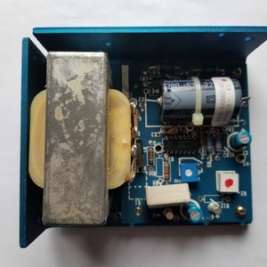 May include: A blue electronic circuit board with various components, including a large transformer, capacitors, resistors, and an integrated circuit. The board is housed in a blue metal enclosure. The components are arranged on a blue surface.