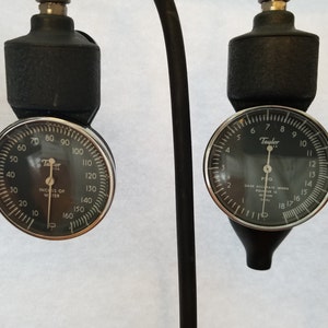 Puede incluir: Dos medidores Taylor negros con bordes plateados. Uno mide en pulgadas de agua, el otro en PSIG. Los medidores tienen caras negras con números y marcas blancas. Los medidores están unidos a tubos negros.