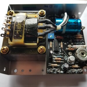 May include: Electronic circuit board with a transformer, capacitors, and various components. The board is mounted in a metal enclosure and secured with gold-colored screws. Wires and other elements are visible.
