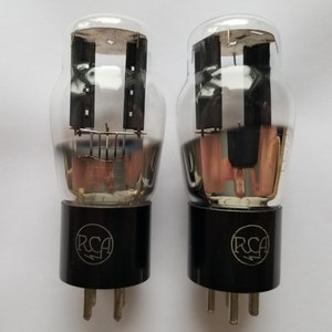 May include: Two vintage RCA vacuum tubes with black bases and clear glass enclosures. The tubes have a copper wire filament and a metal grid inside.