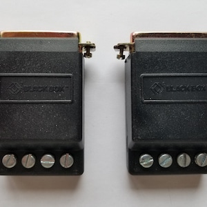 Puede incluir: Dos conectores de plástico negro con conectores de metal dorado y el texto "BLACK BOX" en la parte delantera. Cada conector tiene cuatro tornillos en la parte inferior.