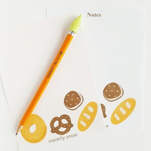May include: Two white notepads with brown and yellow illustrations of bread, pretzels, and bagels. The text "Notes" and "waverly snow" are printed on the notepads. A yellow pencil with a green eraser is lying on top of the notepads.