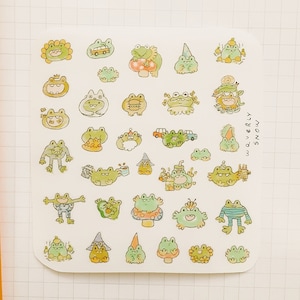 Puede incluir: Una hoja de pegatinas con varias ilustraciones de ranas en diferentes poses y con diferentes accesorios. Las ranas son verdes, amarillas y marrones. Las pegatinas están sobre un fondo blanco.