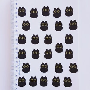 以下が含まれることがあります： 黒いらせん綴じの白いノート。ノートは、黄色い目をした黒い猫のステッカーで覆われています。綴じには「Waverly Snow」という文字が印刷されています。