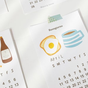 May include: A calendar page for April with a blue and white striped coffee mug and a slice of toast with a fried egg on top. The text "Buongiorno" is at the top of the page.