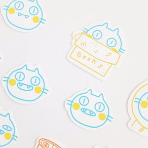 May include: A set of six colorful cat stickers with yellow and blue accents. The stickers feature a cat with a box labeled "BOOKS" and five other cats with different expressions.