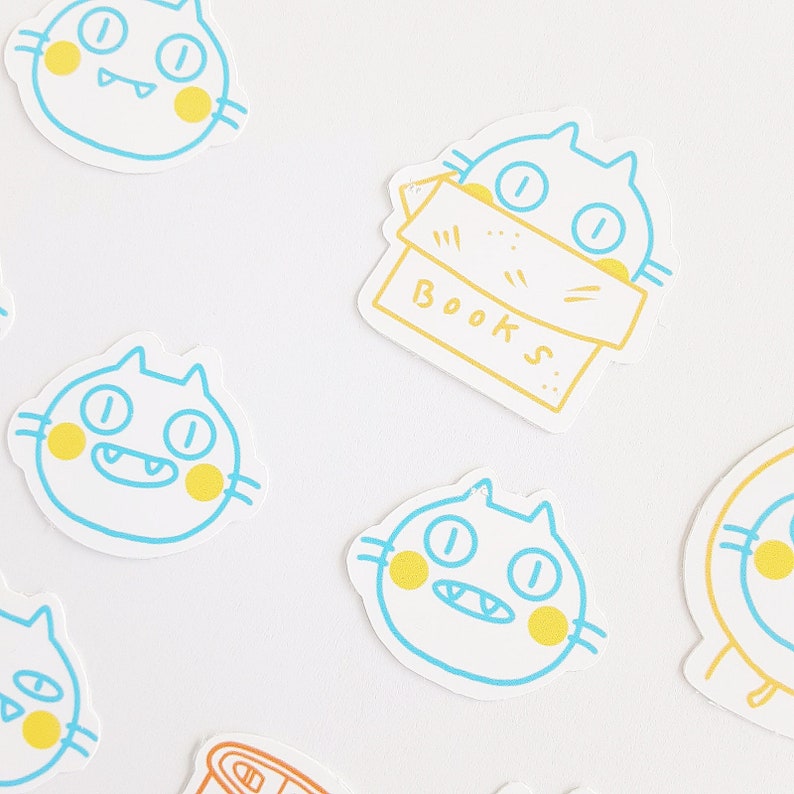 May include: A set of six colorful cat stickers with yellow and blue accents. The stickers feature a cat with a box labeled "BOOKS" and five other cats with different expressions.