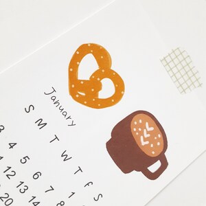 May include: A January calendar page with a pretzel and a cup of coffee. The pretzel is orange and brown with white dots. The coffee cup is brown with white dots and a white swirl on top.