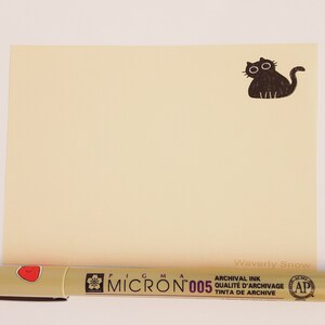 May include: A blank cream-colored card with a black cat illustration in the top right corner. The text "Waverly Snow" is written in black ink at the bottom right corner. A black and beige pen with the text "Pigma Micron 005 Archival Ink Qualite d'Archivage Tinta de Archive" is shown below the card.