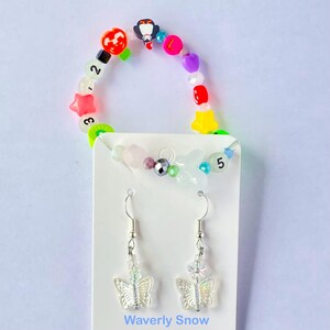 Pode incluir: Uma pulseira de contas coloridas com contas de frutas e números, e um par de brincos de borboleta. Os brincos têm ganchos prateados e pingentes de borboleta iridescentes transparentes. O texto "Waverly Snow" está na parte inferior.