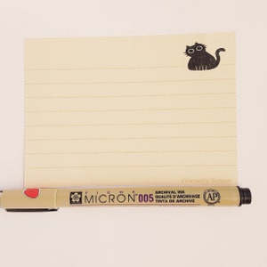 May include: A yellow lined notepad with a black cat illustration and the text "Waverly Snow" at the bottom. A black and gold Micron 005 archival ink pen with a red heart and a pink flower graphic is lying on top of the notepad.