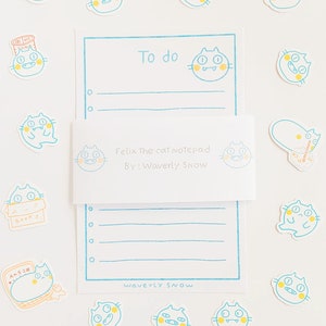 May include: A blue and white notepad with a cat illustration and the text "Felix The Cat Notepad" and "By: Waverly Snow". The notepad has a to-do list section and a lined section. There are several cat stickers around the notepad.