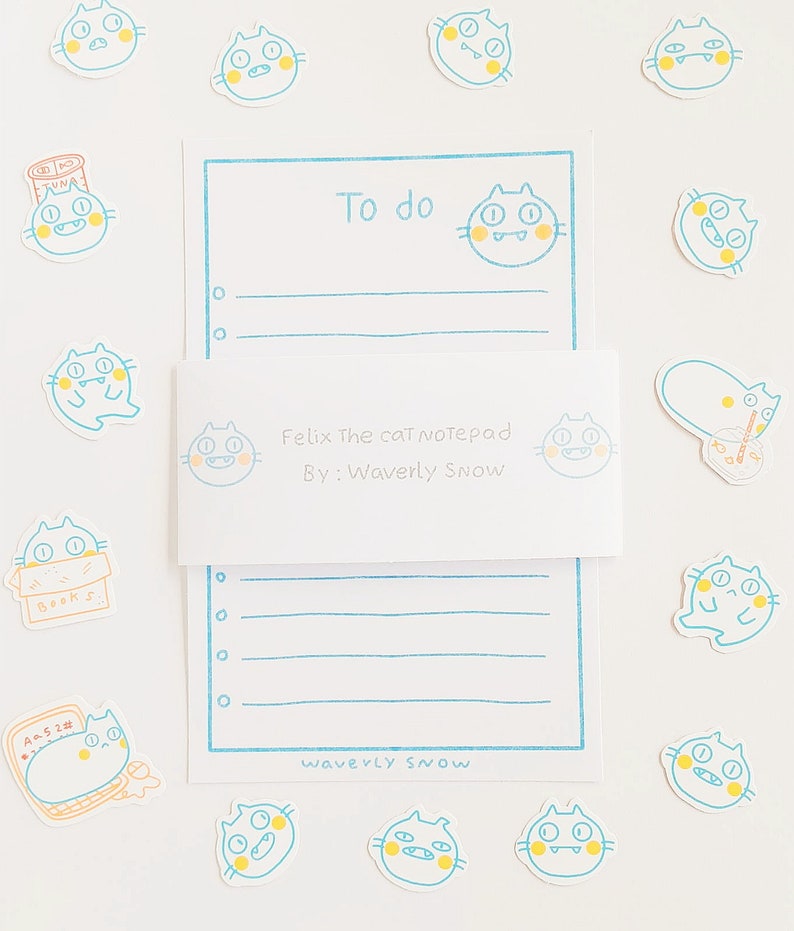 May include: A blue and white notepad with a cat illustration and the text "Felix The Cat Notepad" and "By: Waverly Snow". The notepad has a to-do list section and a lined section. There are several cat stickers around the notepad.