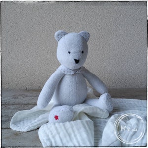 May include: A white plush teddy bear with gray polka dots sits on a white muslin blanket. The bear has a black nose and black eyes. The blanket has a red star on it.