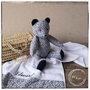 May include: A blue and white floral patterned teddy bear with black ears and a white towel with the name "Edouard" embroidered on it. The bear is sitting on the towel next to a wicker basket.