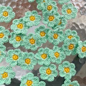 May include: A collection of small, crocheted flower appliqués, each featuring five rounded petals in a light mint green yarn and a bright yellow centre. These individual floral pieces are arranged in a scattered pattern across the frame.