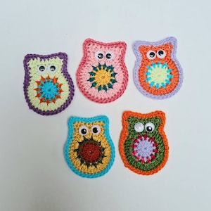 Puede incluir: Cinco apliques de búhos de ganchillo hechos a mano. Cada búho presenta una combinación de colores única, incluyendo morado, rosa, naranja, azul y verde, con círculos interiores contrastantes y ojos saltones. Ideales para proyectos de manualidades.