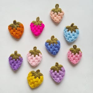 Puede incluir: Una colección de diez pequeñas fresas de ganchillo hechas a mano en varios colores, incluyendo naranja, rosa intenso, melocotón, azul claro, morado, rosa claro, amarillo y lavanda, cada una con un tallo verde oliva oscuro y superficie texturizada.