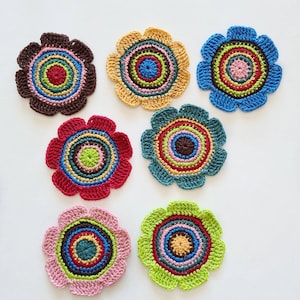 May include: Seven colorful, crocheted flower-shaped coasters. Each coaster features a vibrant, concentric circle design in shades of brown, red, blue, green, yellow, and pink. The coasters are arranged on a white surface.