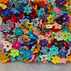 May include: A collection of colorful, crocheted flowers in various shades of blue, pink, yellow, orange, green, and purple. The handmade flowers are clustered together, showcasing their intricate petal designs and textured yarn. The flowers are a mix of sizes and styles.