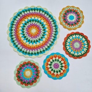 Puede incluir: Cinco tapetes de ganchillo circulares de varios tamaños, cada uno con un vibrante patrón de mandala multicolor con bordes festoneados. Los colores del hilo incluyen tonos de amarillo, naranja, verde, morado, azul y crema en anillos.