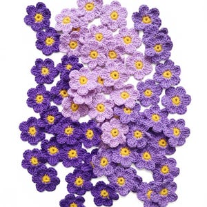 May include: A collection of crocheted flowers in shades of purple and lavender with yellow centers. The handmade floral embellishments are arranged on a white background. These decorative flowers are suitable for various craft projects.