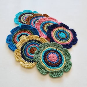 May include: A collection of handmade crochet flower coasters in various colors. Each coaster features a scalloped edge and concentric circles of blue, brown, pink, yellow, and green. The coasters are arranged on a white surface.
