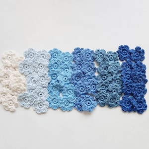 May include: A collection of crocheted flower appliques in a gradient of colors, ranging from off-white to deep blue. The flowers are arranged in rows, showcasing the color variations. The handmade floral elements are ideal for crafting projects.