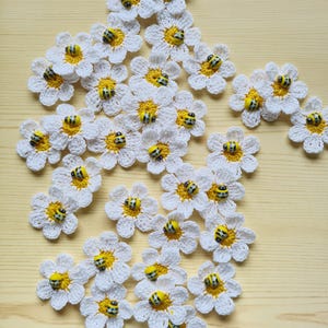 May include: A collection of small, white crocheted daisy appliques, each featuring a bright yellow centre and a tiny, striped bee embellishment. The flowers are arranged in a loose cluster, showcasing the detailed white petals and yellow centres.