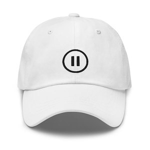 May include: White baseball cap with a black pause symbol embroidered on the front. The cap has a curved brim and an adjustable strap at the back. The hat is made of a soft, breathable fabric.
