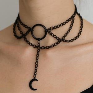 May include: Black choker necklace featuring multiple strands of small chain links. A large circular pendant is centered on the choker, with a chain and crescent moon pendant hanging below. The necklace is worn around the neck.